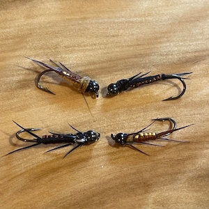 May include: Four fishing flies with curved hooks, designed to mimic insects. Two flies are brown and gold, and two are black. The flies are arranged on a light brown wooden surface.