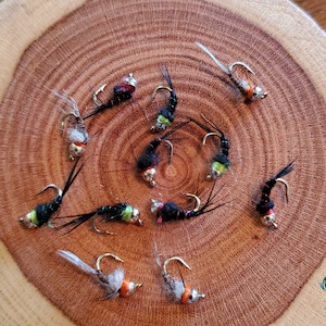 May include: A close-up of ten black, green, and orange fly fishing lures on a wooden surface. The lures have silver hooks and are arranged in a circular pattern.