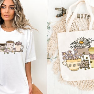 May include: White t-shirt featuring a coffee-themed graphic with cups, flowers, and a spoon. A canvas tote bag displays a similar design with daisies and coffee cups. The design is in soft pastel colors.