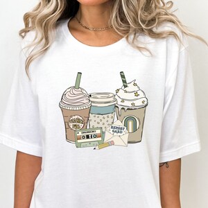 May include: White t-shirt featuring a retro-style graphic with three coffee drinks, a cassette tape, a pencil, and an envelope with the words "Report Card". The coffee cups have whipped cream and star accents.