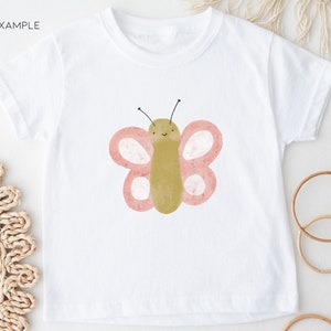 Puede incluir: Camiseta blanca con un gr&aacute;fico de mariposa rosa y verde. La mariposa tiene una cara sonriente y peque&ntilde;as antenas negras.