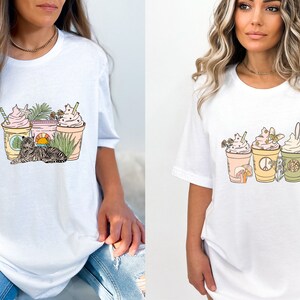 May include: White t-shirts with colorful graphic designs. One shirt features a cat, drinks, and plants. The other shirt has drinks, a mushroom, and a peace sign. The designs are in pastel colors.