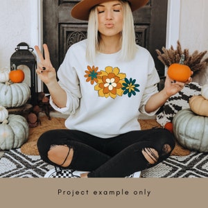 May include: A white sweatshirt with a colourful floral design on the front. The flowers are yellow, orange, green, and brown.