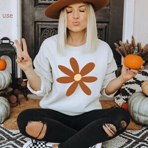 May include: A white sweatshirt with a brown daisy graphic. The daisy has a yellow center. The sweatshirt is being worn by a person sitting on a rug with pumpkins in the background.