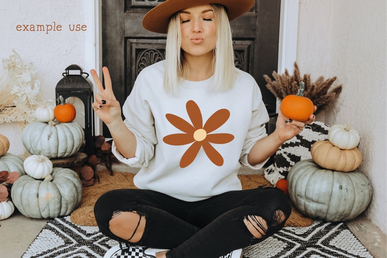 May include: A white sweatshirt with a brown daisy graphic. The daisy has a yellow center. The sweatshirt is being worn by a person sitting on a rug with pumpkins in the background.