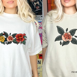 May include: Two white t-shirts with floral designs. The first shirt has a row of three roses with green leaves. The second shirt has a single rose with green leaves.