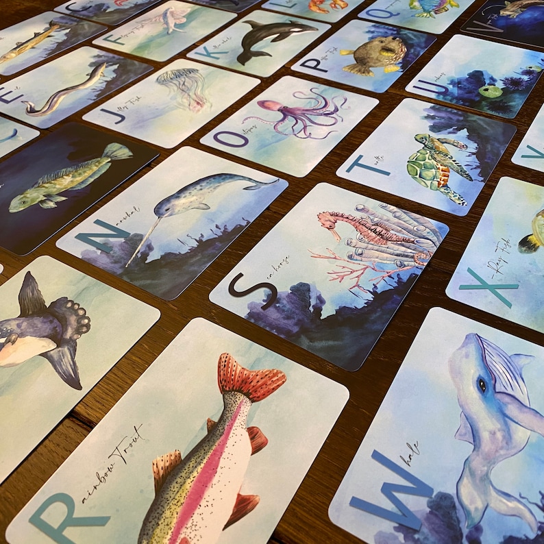 May include: A set of alphabet flash cards featuring colorful illustrations of sea creatures and the corresponding letter. The cards are printed on white cardstock with a blue watercolor background. The letters are in a blue font. The cards include a narwhal, a seahorse, a rainbow trout, a whale, a jellyfish, an octopus, a sea turtle, a starfish, a pufferfish, and more.