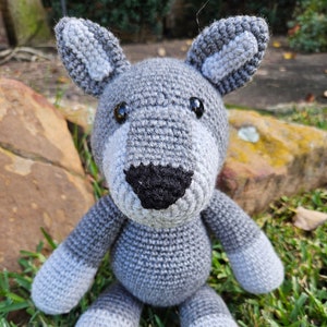 May include: A gray crocheted wolf plush toy with black eyes and a black nose. The toy is sitting on a bed of green grass.
