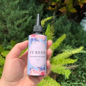 May include: A clear bottle of UV resin for jewelry and craft making. The bottle is 100 grams and has a black cap with a pointed tip. The label is blue with pink flowers and the text "UV Resin Jewelry & Craft Making 100 G".