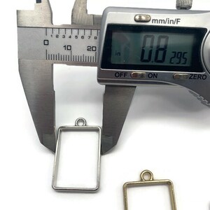 K&ouml;nnte beinhalten: Ein silberner und goldener Metallrahmen-Anh&auml;nger mit einer Messung von 0,8 Zoll, wie auf einer digitalen Schieblehre gezeigt.