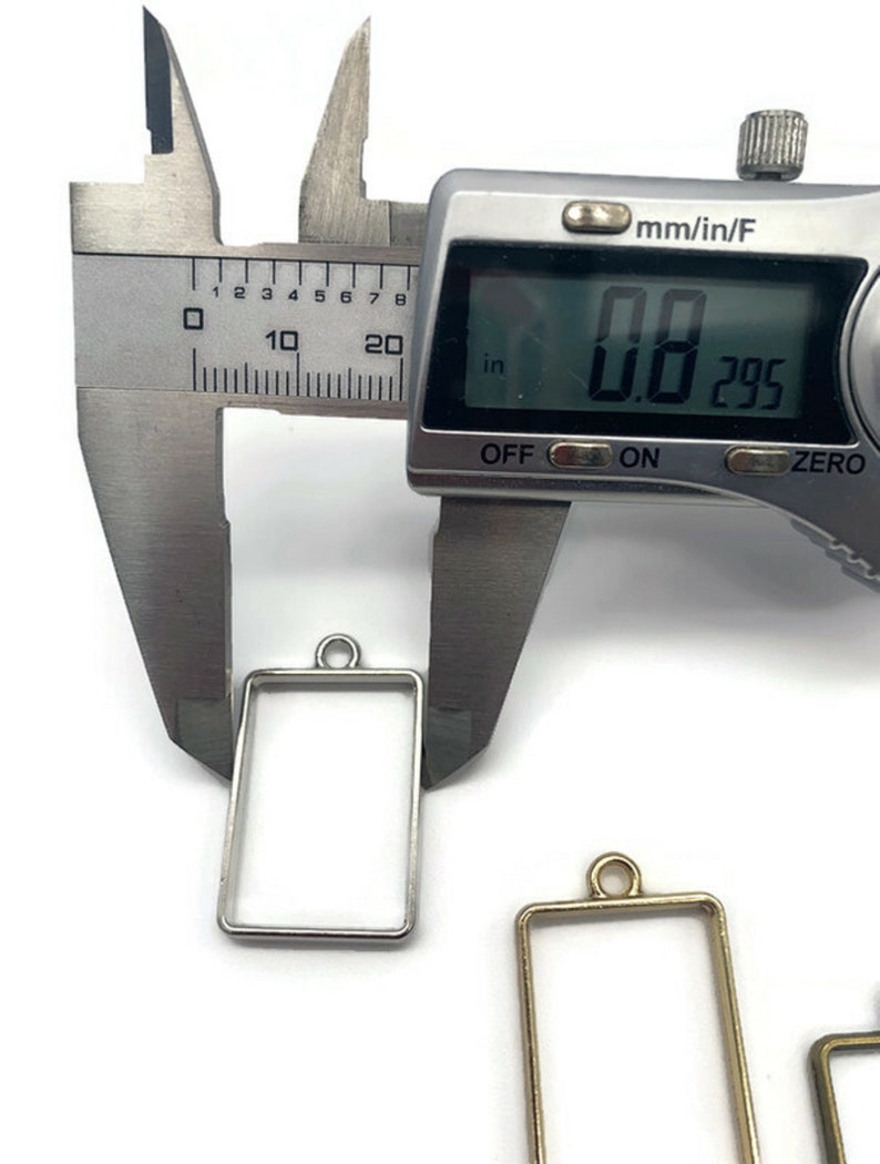 K&ouml;nnte beinhalten: Ein silberner und goldener Metallrahmen-Anh&auml;nger mit einer Messung von 0,8 Zoll, wie auf einer digitalen Schieblehre gezeigt.
