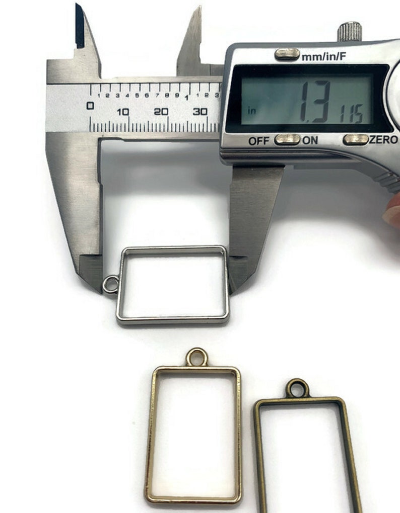 K&ouml;nnte beinhalten: Drei Metallrahmen f&uuml;r Harz-Kunst, einer silber, einer gold und einer bronze. Der silberne Rahmen wird mit einer Schieblehre gemessen, die eine Messung von 3,34 cm anzeigt.