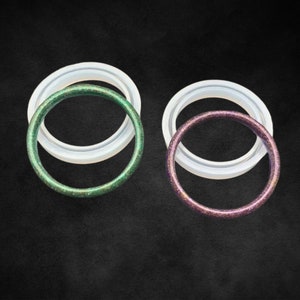 Puede incluir: Dos anillos de resina, uno con acabado de purpurina verde y otro con acabado de purpurina morada. Los anillos están apilados uno encima del otro.