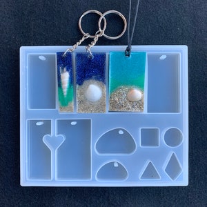 May include: A clear silicone mold with multiple shapes for making resin jewelry. The mold includes rectangular, heart, oval, square, circle, and triangle shapes. Three rectangular pieces of resin jewelry with seashells and glitter are shown in the mold.