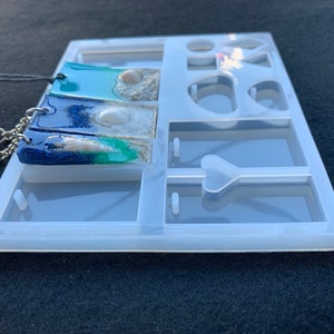 May include: A clear silicone mold for making resin jewelry. The mold has multiple compartments, including two rectangular shapes for pendants, a heart shape, and several round shapes. The mold is shown with two finished resin pendants, one with a blue and green color scheme and the other with a blue and white color scheme.