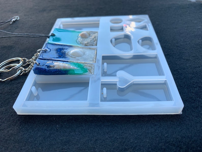 May include: A clear silicone mold for making resin jewelry. The mold has multiple compartments, including two rectangular shapes for pendants, a heart shape, and several round shapes. The mold is shown with two finished resin pendants, one with a blue and green color scheme and the other with a blue and white color scheme.
