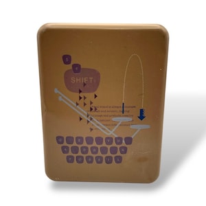 May include: A rectangular, tan-colored box with a design featuring the word "SHIFT" and a keyboard layout. The design includes arrows and text, with the brand name "FOSSIL" spelled out on the keyboard.