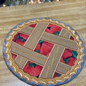 May include: A round, fabric coaster with a strawberry pie design. The center features a fabric print of red strawberries. Tan fabric strips form a lattice pattern, stitched with gold thread. The edge is a scalloped design in gold and blue.