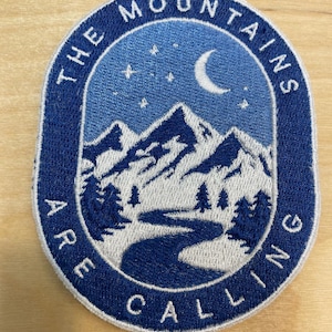 May include: A blue and white embroidered patch with the text "THE MOUNTAINS ARE CALLING" in a circle. The patch features a mountain range with a river, trees, and a crescent moon with stars.