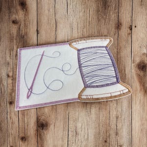 May include: A white felt sewing themed coaster with a purple thread spool and a pink needle with a thread loop. The coaster has a light brown border.