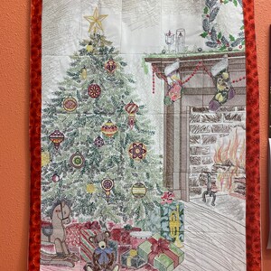 May include: A Christmas-themed wall hanging featuring a decorated tree, fireplace, and wrapped gifts. The tree is adorned with ornaments and topped with a star. The border is a dark red with a patterned design. A teddy bear and rocking horse are visible.