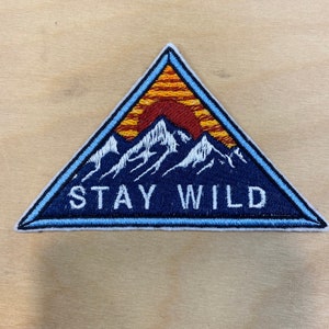 May include: A blue triangular patch with a white mountain range and a red and orange sunset. The text "STAY WILD" is embroidered in white on the patch.