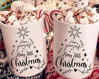 May include: Two white mugs filled with hot chocolate and marshmallows. The mugs have black text that reads "Have yourself a Merry Little Christmas". The mugs are sitting on a blanket with a red and white pattern.