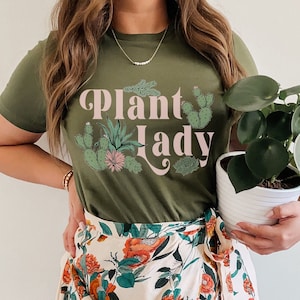 May include: A green t-shirt with the text "Plant Lady" in pink letters and a graphic of cacti and succulents. The person wearing the shirt is holding a potted plant.