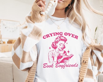 May include: White t-shirt with a pink graphic of a woman reading a book and the text "Crying Over Book Boyfriends".