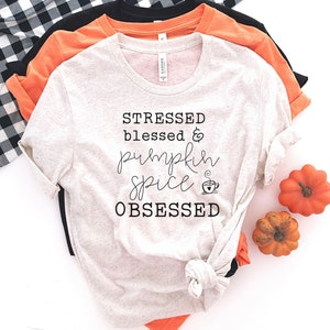 May include: A beige t-shirt with the text "Stressed Blessed & Pumpkin Spice Obsessed" and a small coffee cup graphic. The shirt is folded and displayed on a white background with two small orange pumpkins.
