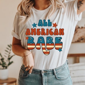 May include: A white t-shirt with a red, white and blue graphic that says "All American Babe" in a retro font.