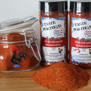 May include: Two glass jars of Tia Maria's Portuguese Seasoning, one labeled "Spicy". The jars are filled with a red-orange powder and have a rooster logo on the front. The jars are sitting on a wooden surface with a pile of the seasoning in front of them.