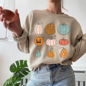 May include: Beige sweatshirt featuring a colorful pumpkin design. The pumpkins vary in color and pattern, including stars, leopard print, and stripes. The design is perfect for fall and Halloween.