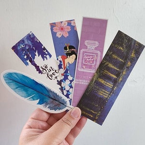 May include: Five different bookmarks with various designs. One bookmark features a blue watercolour feather with the text "So in love". Another bookmark has a Japanese woman in a blue kimono with pink cherry blossoms. A third bookmark has a purple background with a neon pink outline of a perfume bottle with the text "Space Jolie" and the date "12 December". The fourth bookmark has a blue and gold abstract design. The fifth bookmark has a blue and white watercolour design.