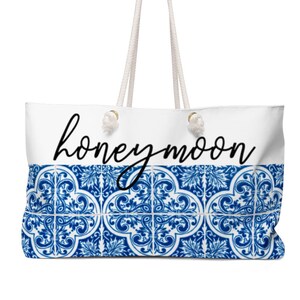 Op de afbeelding: Witte draagtas met touwhandvatten en een blauw-wit tegelpatroon aan de onderkant. Het woord "honeymoon" staat in zwart script over de bovenkant van de tas.