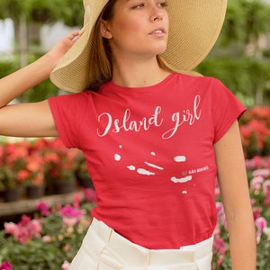May include: A woman wearing a red t-shirt with the text "Island girl" and a white outline of an island with the text "540 miles" printed on it. She is wearing a straw hat and white trousers.