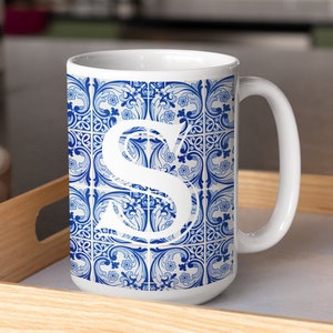 May include: White ceramic mug with a blue and white patterned design featuring the letter "S". The mug is on a wooden tray with a glass next to it.