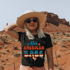 May include: A woman wearing a black t-shirt with the text "All American Babe" in red, white and blue. She is wearing a white hat and sunglasses and is standing in front of a red rock formation.