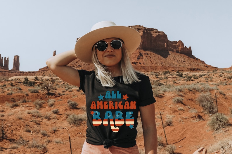 May include: A woman wearing a black t-shirt with the text "All American Babe" in red, white and blue. She is wearing a white hat and sunglasses and is standing in front of a red rock formation.