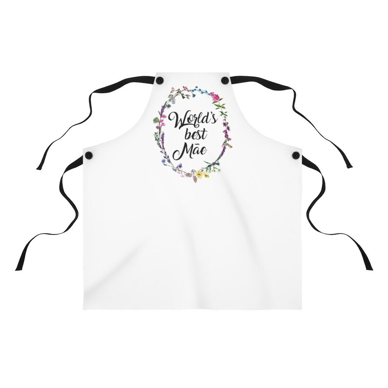 May include: White apron with black straps and a floral wreath design. The wreath has the text "World's best M&atilde;e" in black script.