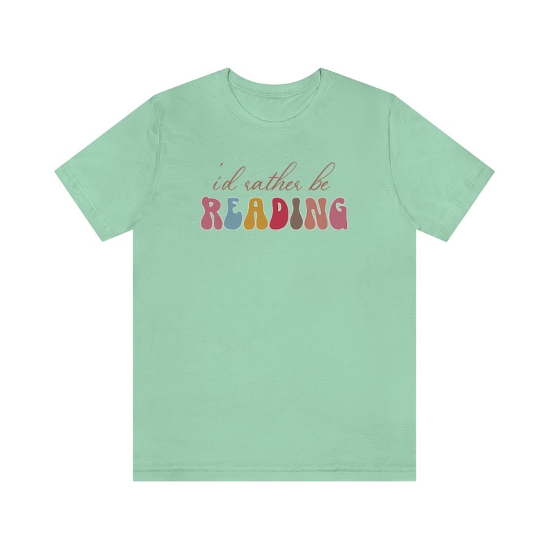 Puede incluir: Una camiseta verde claro con el texto "i'd rather be READING" en letras de colores de estilo retro.