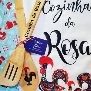 May include: A white kitchen towel with the text "Cozinha da Rosa" in black cursive lettering. Two wooden spoons with a rooster design are on top of the towel. One spoon has the text "Cozinha da Rosa" engraved on it. A small blue tag with the text "Lisbon Blue" is attached to the spoons.