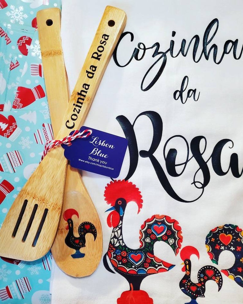 May include: A white kitchen towel with the text "Cozinha da Rosa" in black cursive lettering. Two wooden spoons with a rooster design are on top of the towel. One spoon has the text "Cozinha da Rosa" engraved on it. A small blue tag with the text "Lisbon Blue" is attached to the spoons.