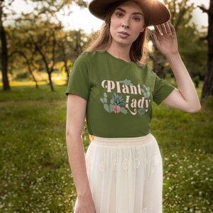 May include: A woman wearing a green t-shirt with the text "Plant Lady" in pink lettering and a white skirt. She is wearing a brown hat and is standing in a field of green grass.