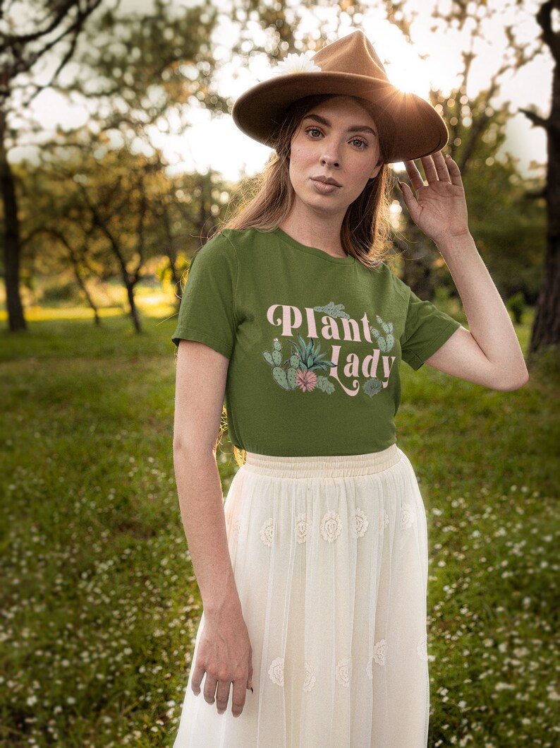 May include: A woman wearing a green t-shirt with the text "Plant Lady" in pink lettering and a white skirt. She is wearing a brown hat and is standing in a field of green grass.