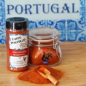 May include: A glass jar and a plastic bottle of "Taste Portugal Tia Maria's Portuguese Seasoning" with a red rooster logo. The seasoning is red and is spilling out of the jar onto a wooden surface. The jar has a metal clasp.