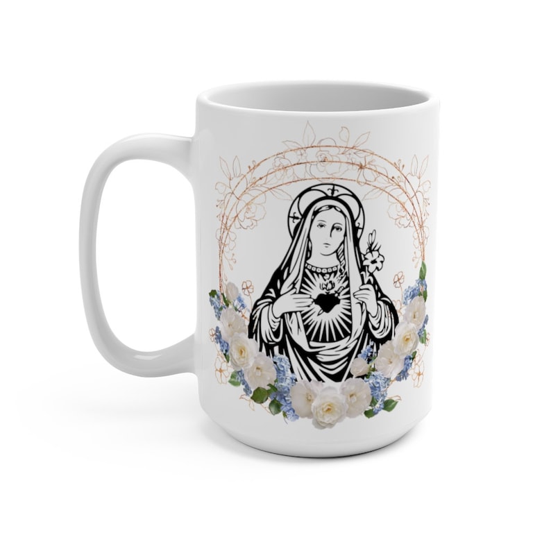 May include: White ceramic mug with a black and white image of the Virgin Mary with a heart in her chest. The image is surrounded by a wreath of white roses and blue flowers with gold accents.