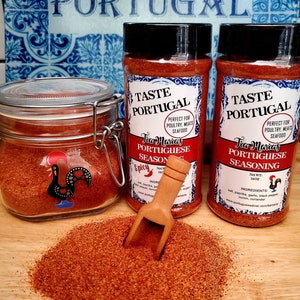 May include: Two glass jars of Tia Maria's Portuguese Seasoning, one open and one closed. The jars are labeled "Taste Portugal" and "Perfect for Poultry, Meats, Seafood". The seasoning is a red-brown color and is piled on a wooden surface.