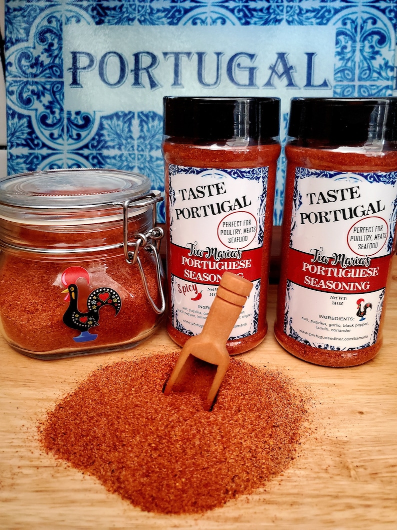 May include: Two glass jars of Tia Maria's Portuguese Seasoning, one open and one closed. The jars are labeled "Taste Portugal" and "Perfect for Poultry, Meats, Seafood". The seasoning is a red-brown color and is piled on a wooden surface.