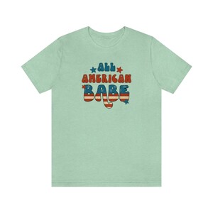 May include: A light green t-shirt with the text "All American Babe" in a retro font with red, white and blue stars and stripes.